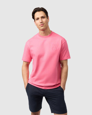 A man wearing a pink Psycho Bunny Pima cotton t-shirt with a small embroidered appliqué bunny logo and black shorts standing against a plain light background. He appears neat with short dark hair and a serious expression.