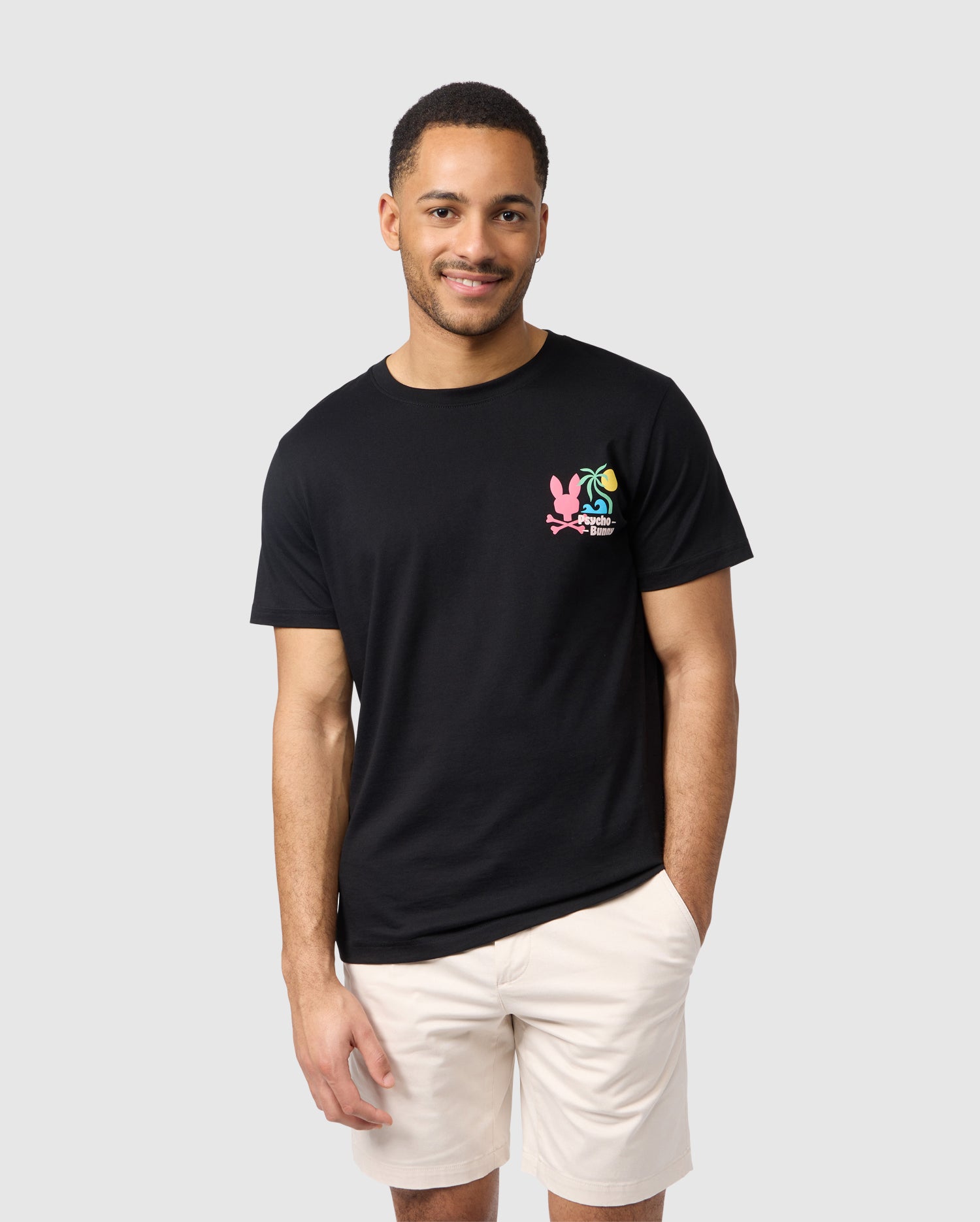 A man with short hair smiles at the camera, wearing a black Psycho Bunny MENS MENTZ GRAPHIC TEE - B6U395B200 featuring a colorful tropical-inspired logo on the left chest and light-colored shorts. His left hand is in his pocket as he stands against a plain, light grey background.