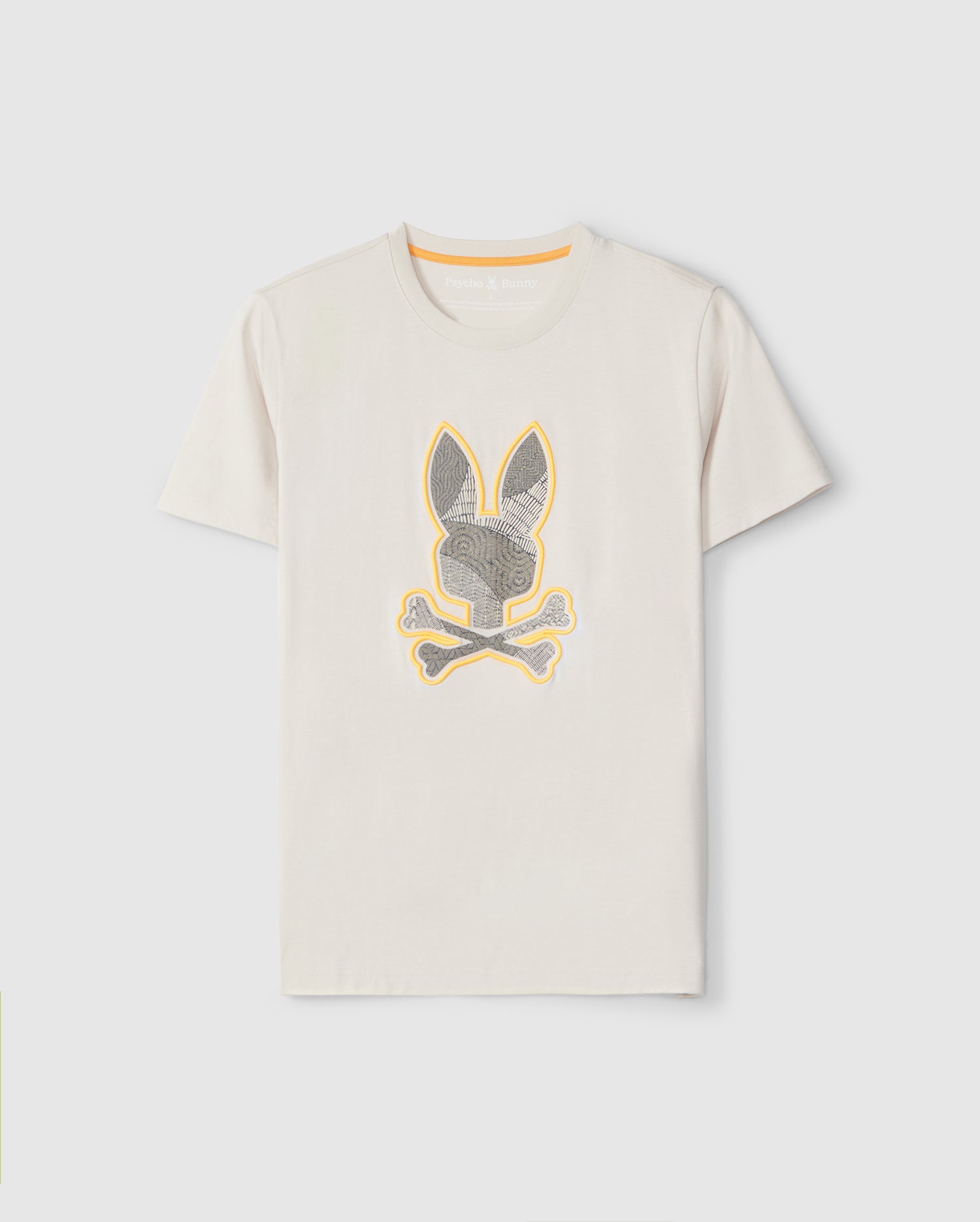 A plain light beige Pima cotton tee featuring an embroidered print on the front. The design includes a stylized bunny head with long ears and intricate patterns, placed above two crossed bones. The artwork is outlined in yellow, contrasting with the shirt's neutral tone. This is the MENS LENOX EMBROIDERED GRAPHIC TEE - B6U405B200 by Psycho Bunny.