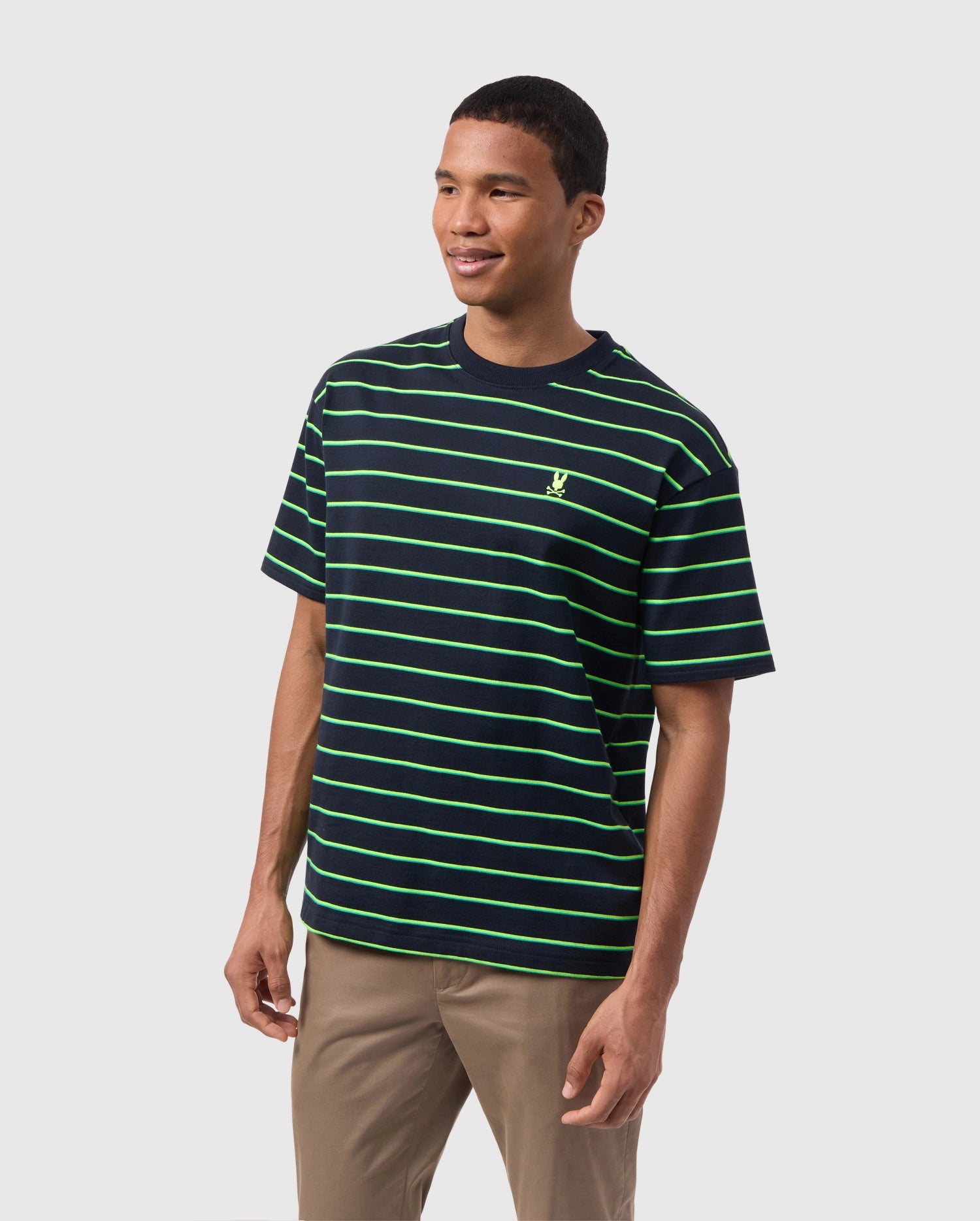 A person is standing and smiling, wearing a black Psycho Bunny MENS ALTON STRIPE OVERSIZED TEE - B6U409C200 with 90s-inspired two-tone stripes and beige pants. The relaxed fit complements the light-colored background, adding to the casual yet stylish look.