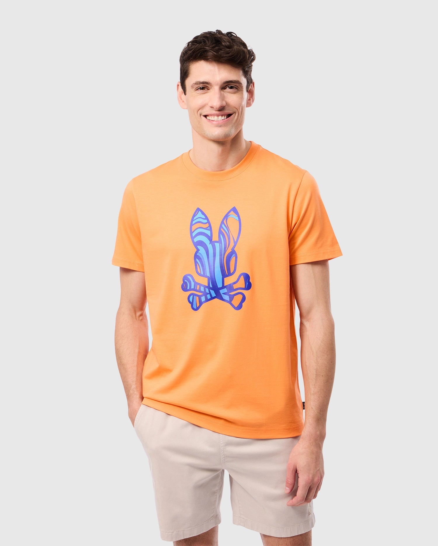 A man wearing a Psycho Bunny MENS NEVADA GRAPHIC TEE - B6U515C200 featuring a blue bunny print on the front stands against a plain light gray background. Made of soft Pima cotton, he is smiling with one hand in the pocket of his light beige shorts.