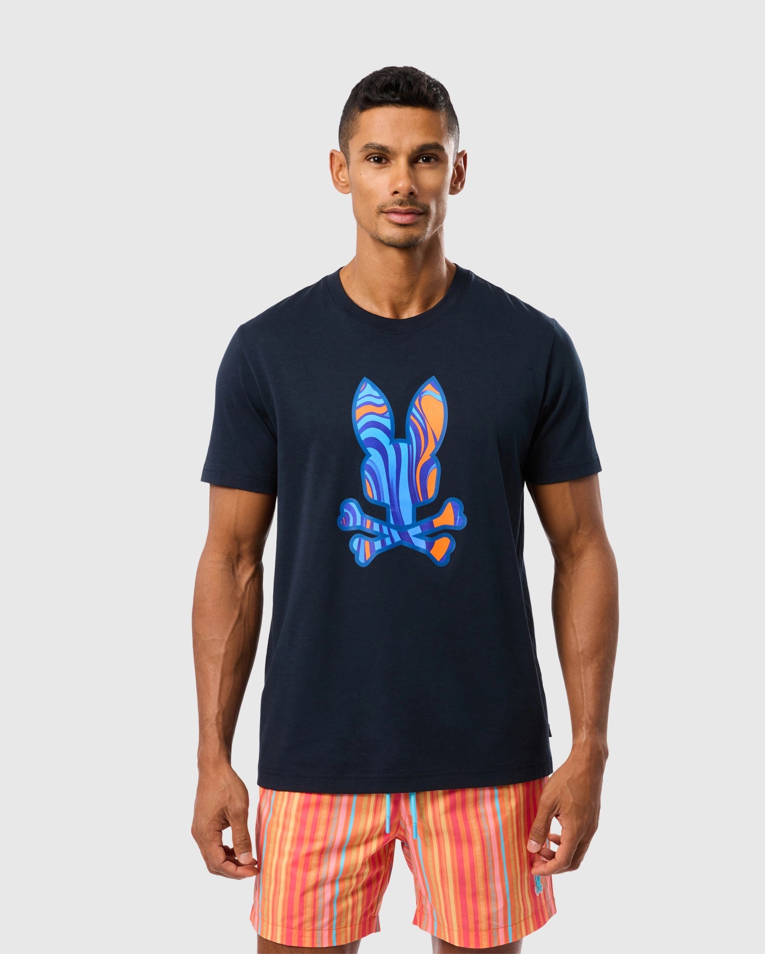 A man is wearing a navy blue MENS NEVADA GRAPHIC TEE - B6U515C200 by Psycho Bunny, crafted from soft Pima cotton and showcasing a vibrant, abstract design of two surfboards and crossed paddles on the front. Additionally, he is sporting colorful striped shorts in shades of orange and pink against a plain white background.