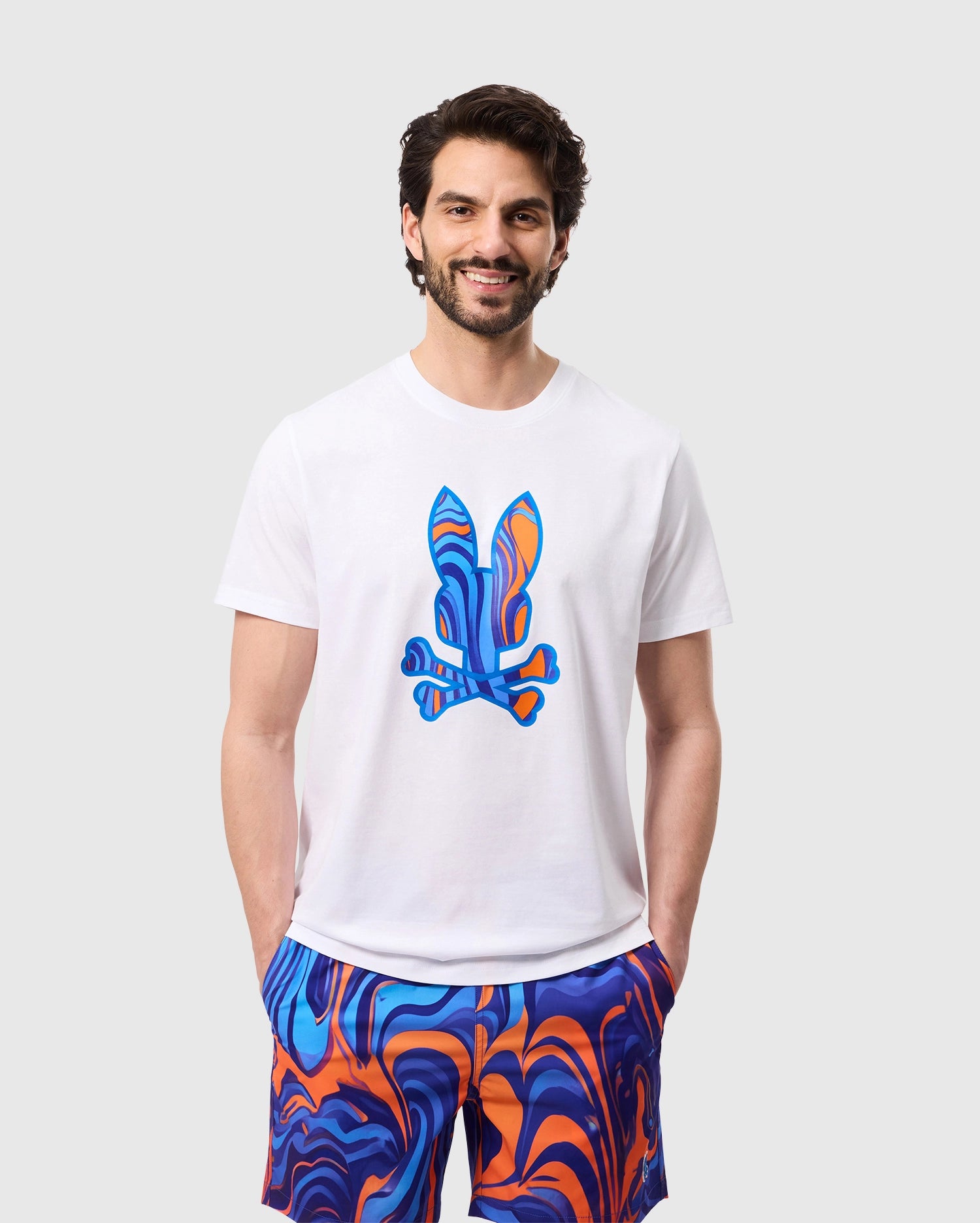 A man with a beard is smiling, wearing a white MENS NEVADA GRAPHIC TEE - B6U515C200 by Psycho Bunny made from soft Pima cotton, featuring a colorful abstract bunny graphic on the front. He pairs it with matching bright blue and orange swirl-patterned shorts. He has his hands in his pockets and is standing against a plain light gray background.