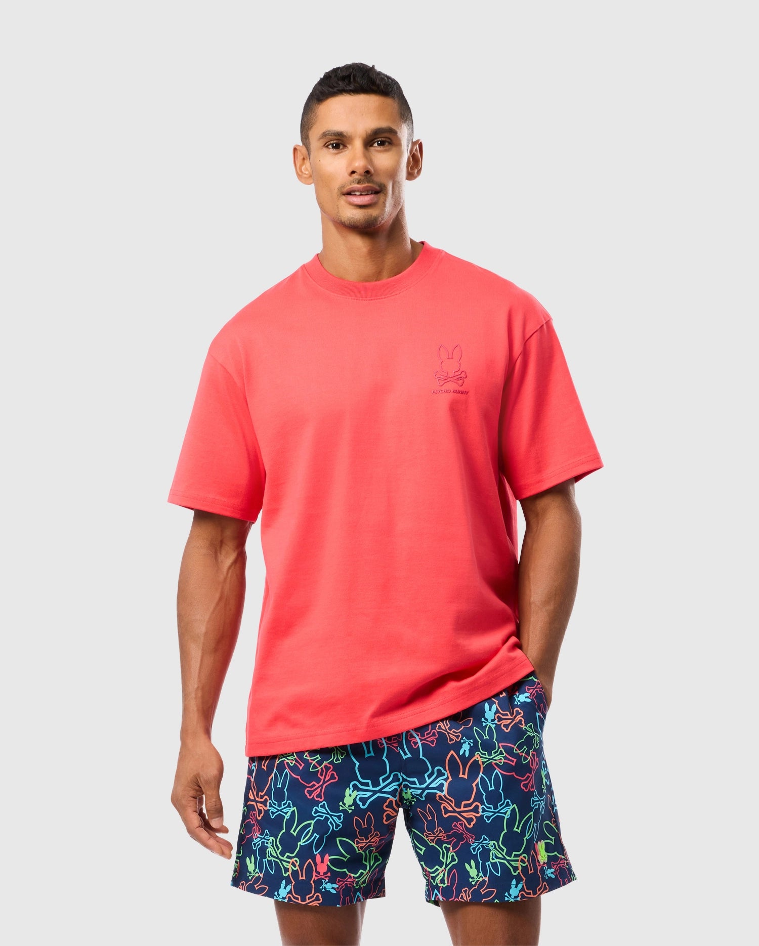 A man stands against a plain background wearing a bright red T-shirt in relaxed fit and colorful shorts with neon animal patterns. He has short dark hair and a neutral expression. The Psycho Bunny MENS BARRETT RELAXED FIT TEE - B6U518C200 features a small embroidered bunny graphic on the left chest area.