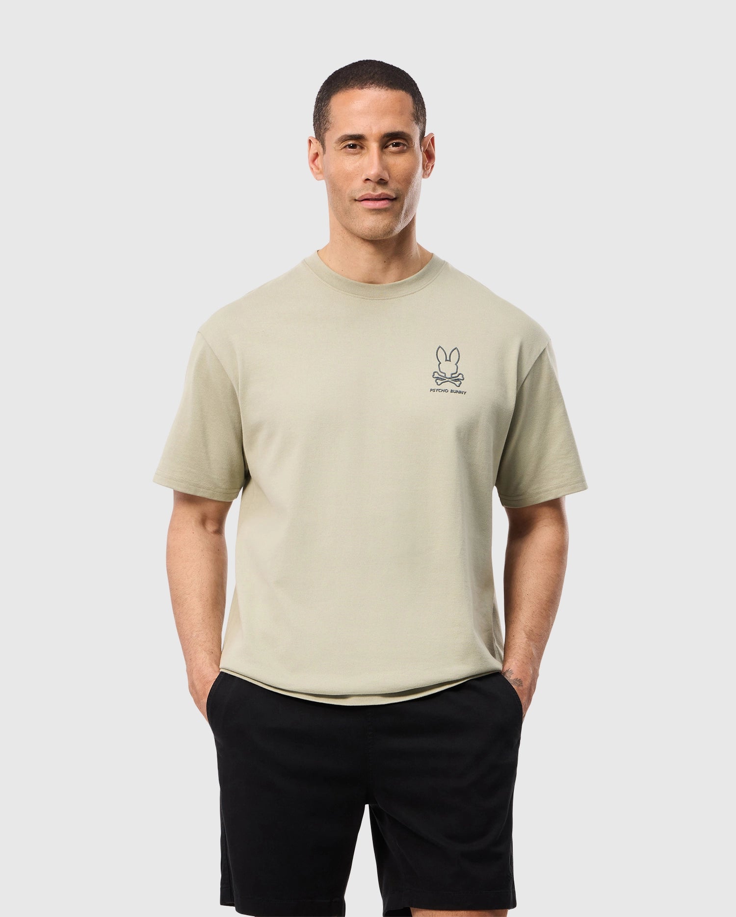A man stands confidently with his hands in his pockets, wearing a Psycho Bunny MENS BARRETT RELAXED FIT TEE - B6U518C200, a light beige T-shirt made of heavyweight Pima cotton, featuring a small Bunny graphic above his left chest and black shorts. He has short hair and a neutral expression, posed against a plain light grey background.