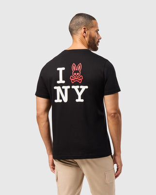 A man with short hair and a beard is wearing a Psycho Bunny MENS NEW YORK TEE - B6U554W1PC in black, featuring "I NY" in white letters and a red bunny-skull graphic on the back. Made from Pima cotton, the shirt drapes well against his beige pants as he faces slightly away from the camera, showcasing the back design.