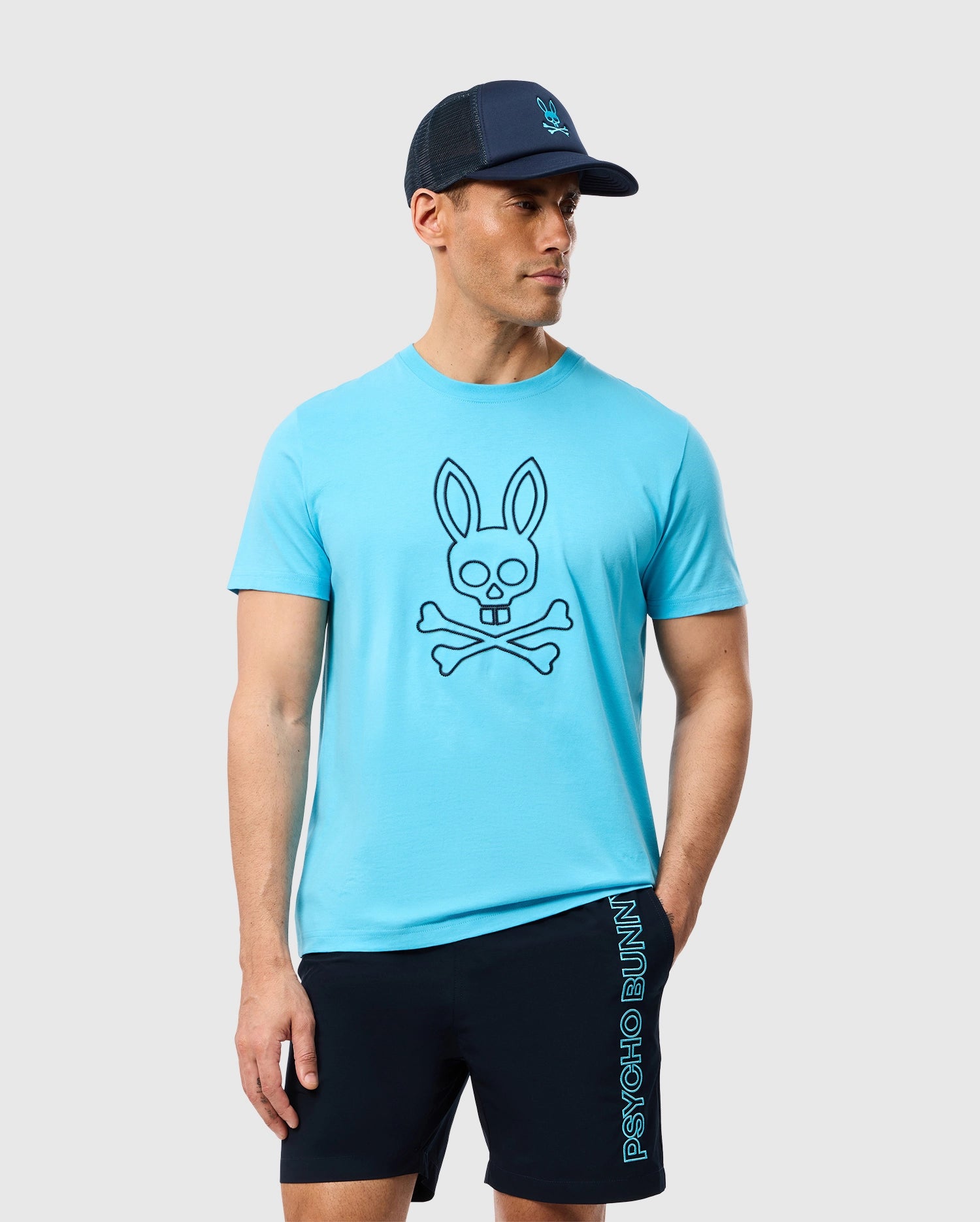 A man wearing the Psycho Bunny MENS SHELDON GRAPHIC TEE - B6U569C200 in turquoise, featuring a skull and crossbones with a bunny head. He is also in navy shorts with 