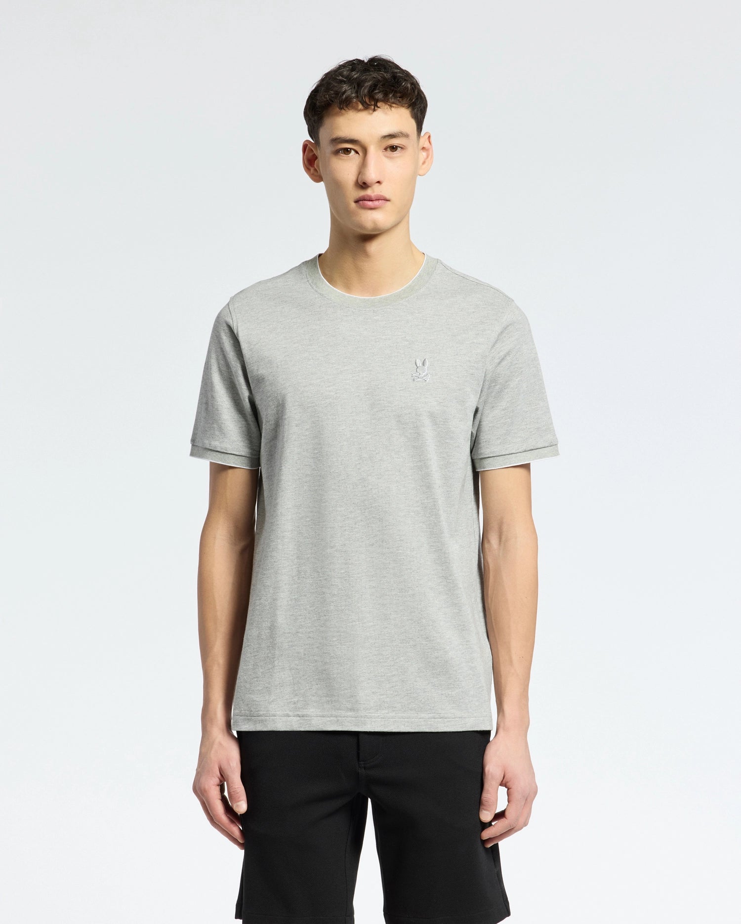 A person with short dark hair is wearing a light gray Psycho Bunny MENS MACKAY JACQUARD FASHION TEE - B6U576F200 with an embroidered Bunny logo on the upper left chest, paired with black shorts, against a plain white background.