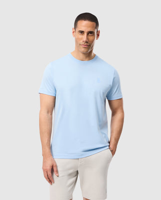 A man stands against a plain background, wearing a light blue Psycho Bunny MENS MASON GRAPHIC TEE - B6U610C200 and beige shorts. His hands are relaxed at his sides, highlighting the subtle elegance of this comfortable back print tee.