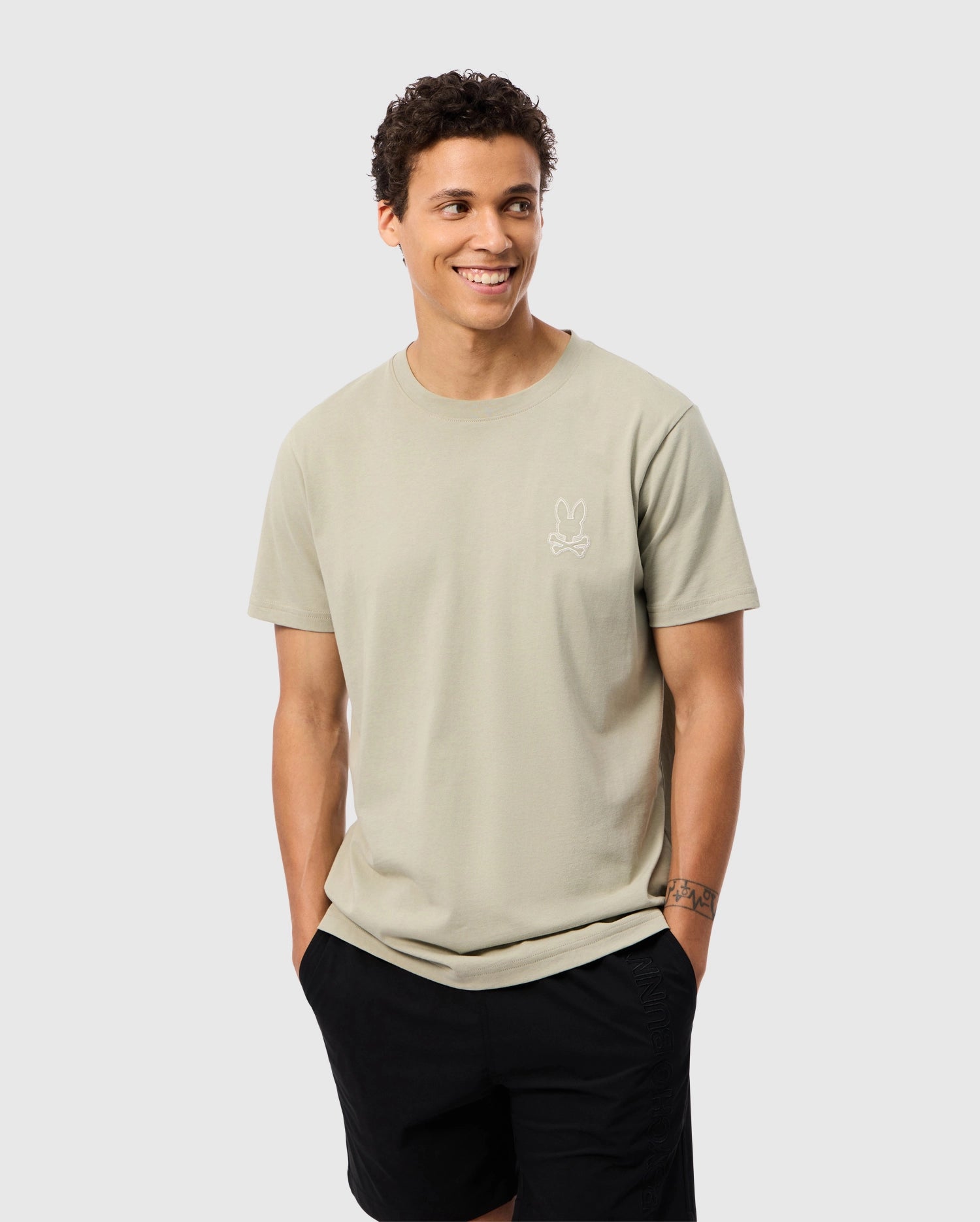 A smiling person with short curly hair stands against a plain background, wearing the luxurious light beige Psycho Bunny Men's Pearland Back Graphic Tee (B6U632C200) made from Pima cotton, featuring a small minimalist bunny emblem embroidered on the left side of their chest, and black shorts. Their hands are in their pockets.