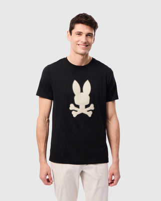 A person with short dark hair is standing and smiling. They are wearing a black Psycho Bunny MENS RIVIERA GRAPHIC TEE - B6U639C200 featuring a silicon HD design of a rabbit head above crossbones, and light-colored pants. The background is plain white.