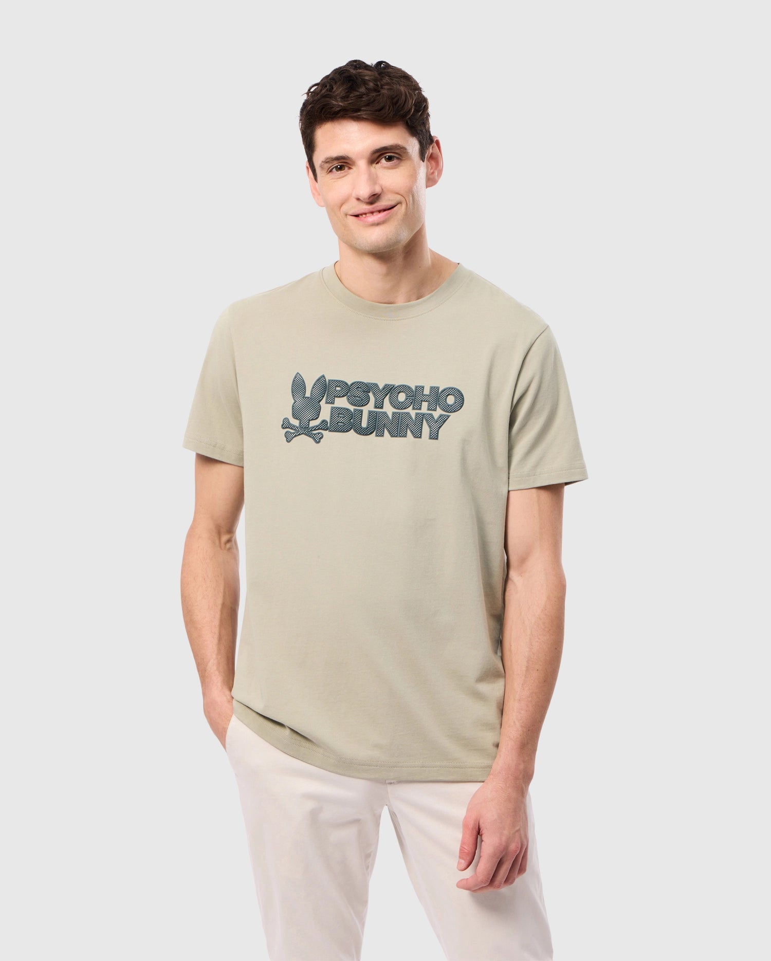 A man with short, dark hair is smiling and posing, standing with one hand in his pocket. He is wearing a light green MENS MELVIN GRAPHIC TEE - B6U663C200 by Psycho Bunny with the words 