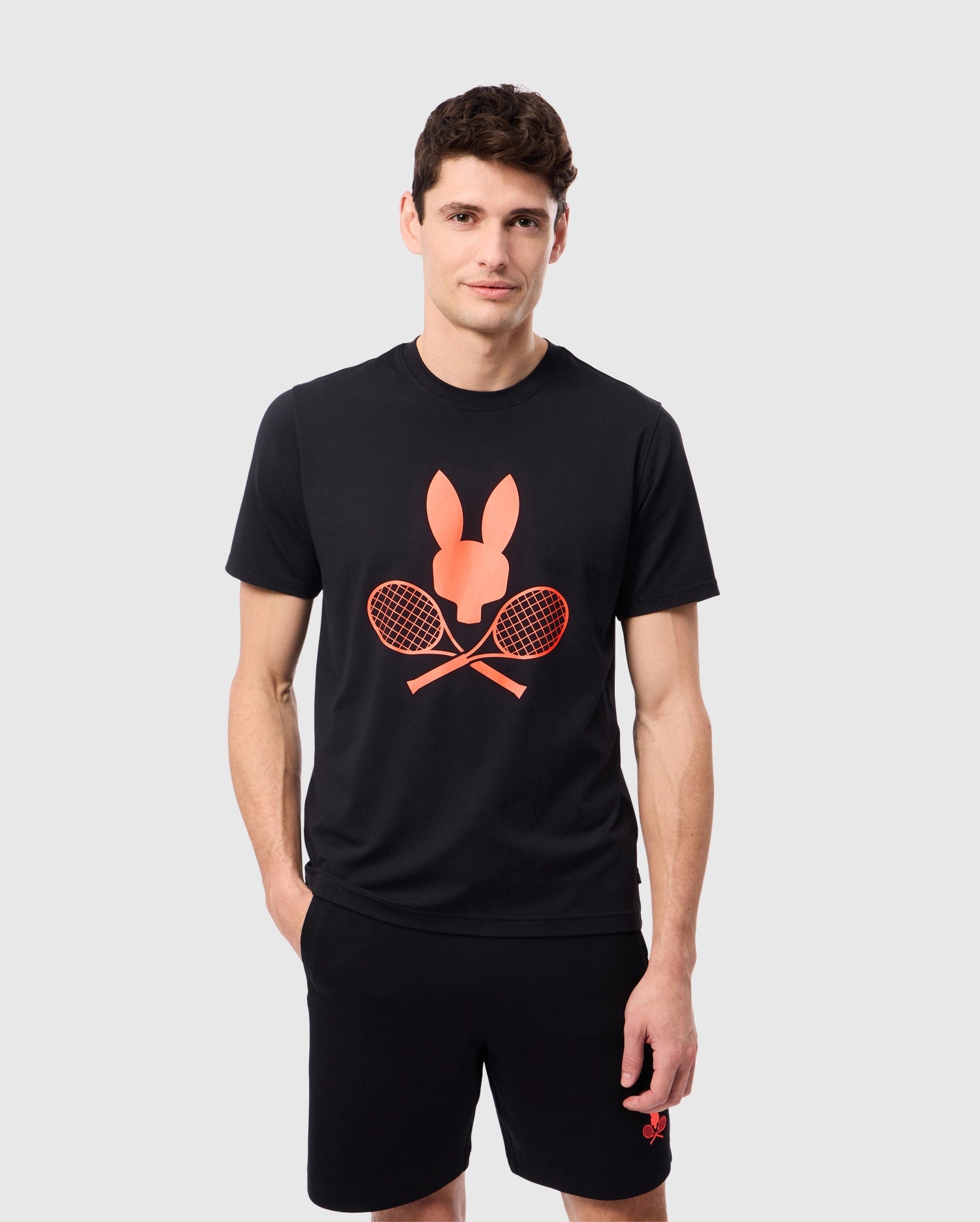 A man is modeling a Psycho Bunny MENS COURTSIDE GRAPHIC TEE - B6U681C200, which showcases a red tennis-themed Bunny logo with crossed rackets. With short brown hair, he strikes a pose against a simple light gray backdrop and pairs the tee with black shorts, gazing directly into the camera.