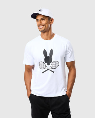 A man wearing a white cap and a white Psycho Bunny MENS COURTSIDE GRAPHIC TEE - B6U681C200 with a black tennis-inspired Bunny logo on the front. He is smiling, with his hands in the pockets of his black pants, standing against a plain light gray background.