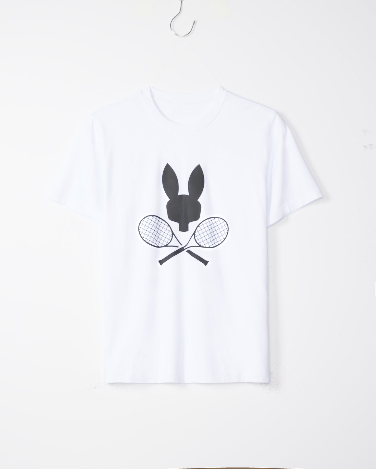 A MENS COURTSIDE GRAPHIC TEE - B6U681C200 is hanging on a wall hook. The white shirt features a black silhouette of a tennis-inspired Bunny logo above two crossed racquets in the center. The background is plain and minimalistic. This stylish tee is from Psycho Bunny.