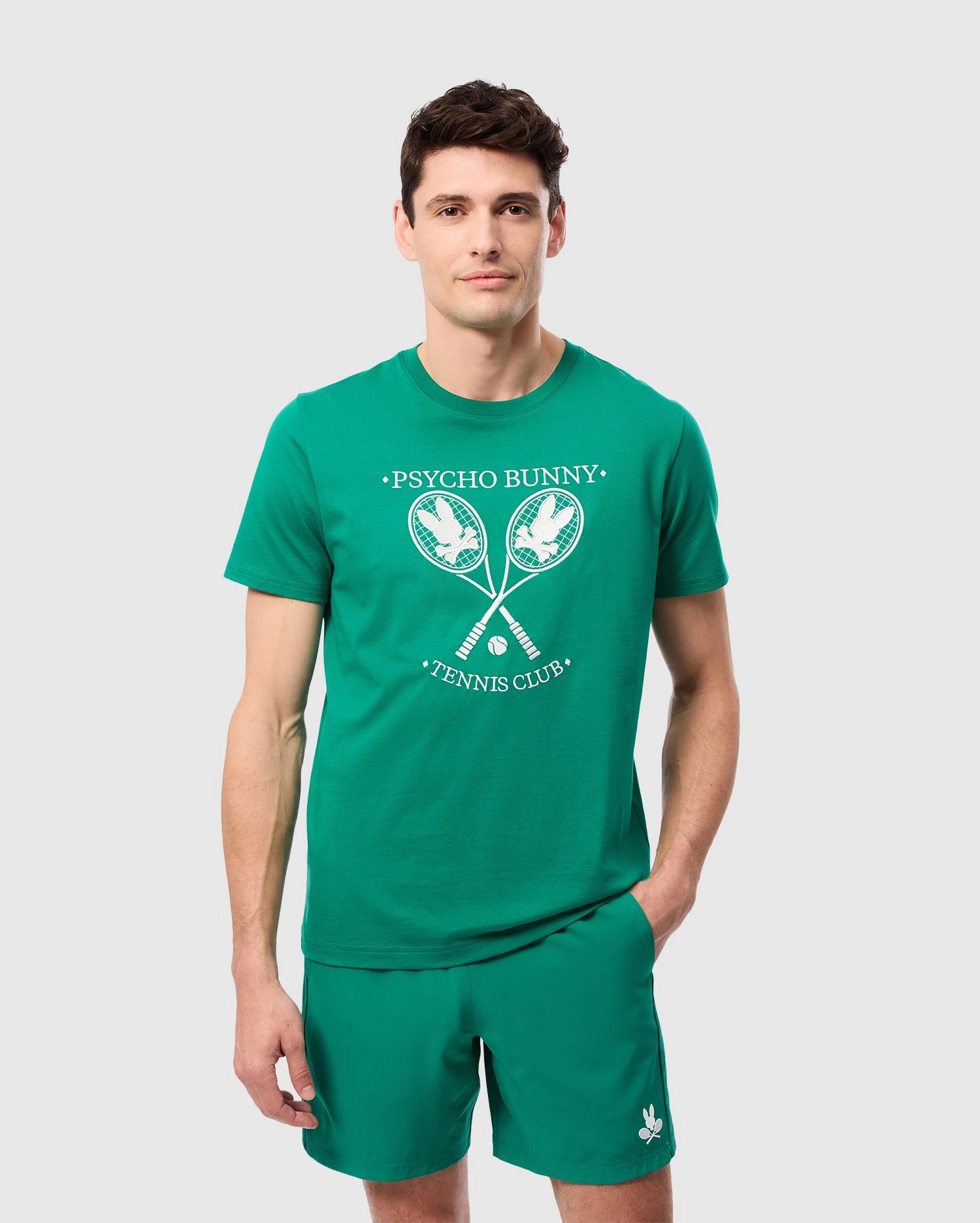 A man in a green MENS COURTSIDE GRAPHIC TEE - B6U684C200 and green shorts stands against a plain white background. The t-shirt, crafted from Pima cotton jersey, features a tennis-inspired Bunny logo with two crossed rackets and 