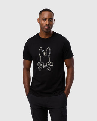 A man wearing the Psycho Bunny MENS VINCENT GRAPHIC TEE - B6U713D200, which features a rabbit head above crossed bones. Made from durable Pima cotton, the black t-shirt complements his short hair and neutral expression as he stands against a plain white background with his hands in his pockets.