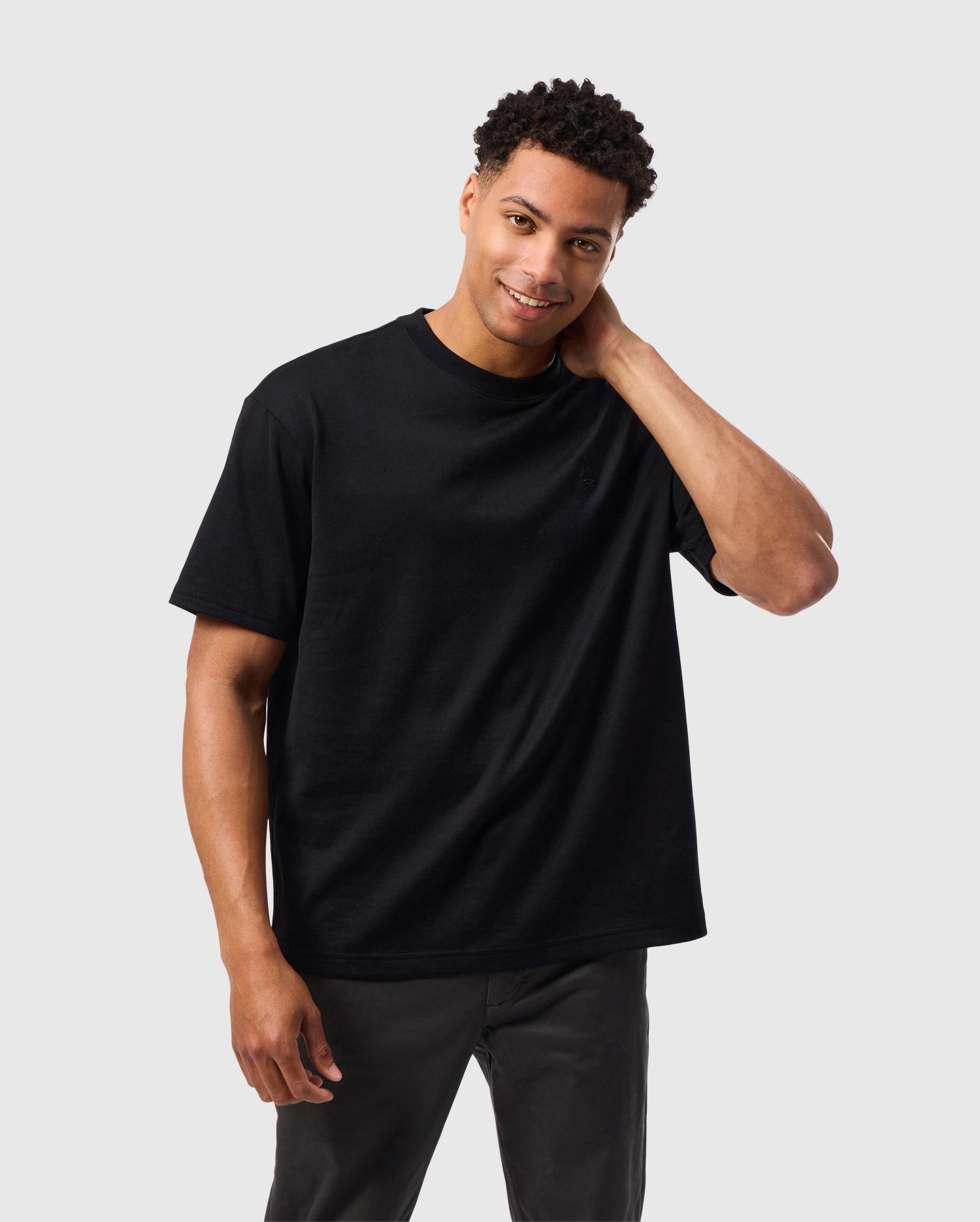 A man with short, curly hair is smiling and looking at the camera. He is standing against a plain, light gray background, wearing a relaxed fit Psycho Bunny MENS DUKE OVERSIZED TEE - B6U742D200 and black pants. His left hand is touching the back of his neck.