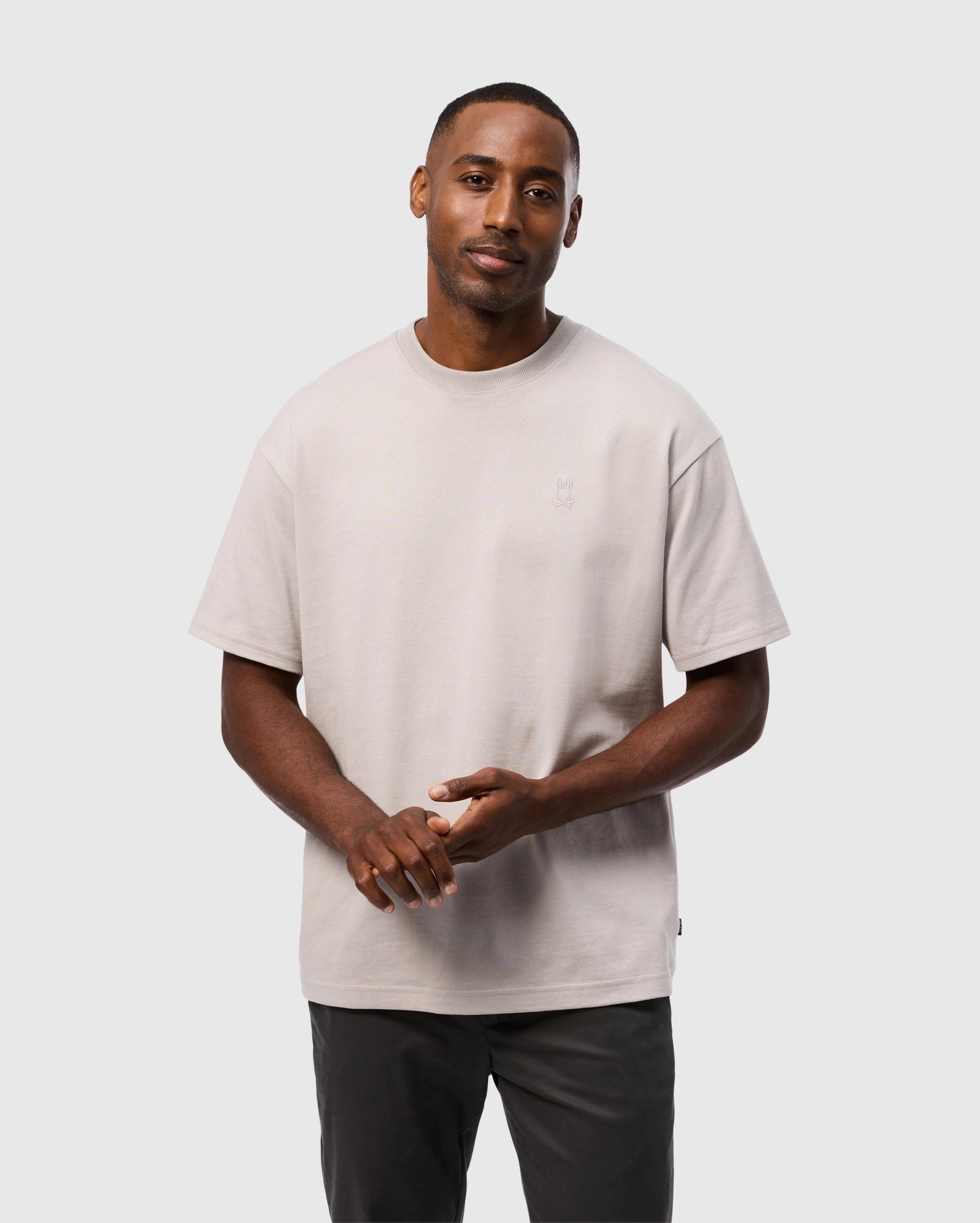 A man wearing the Psycho Bunny MEN'S DUKE OVERSIZED TEE - B6U742D200 in light grey, a relaxed fit made of heavyweight Pima cotton, and dark pants stands against a plain white background. His hands are loosely clasped in front of him, and he is looking towards the camera with a neutral expression.