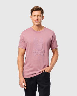 A person wearing the Psycho Bunny MENS KINGSTON GRAPHIC TEE - B6U743D200 in pink, with a discreet embossed bunny and crossbones design, stands against a plain light gray background, one hand tucked into the pocket of their black pants.