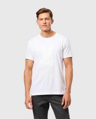 A man stands against a plain backdrop wearing a Psycho Bunny MENS KINGSTON GRAPHIC TEE - B6U743D200 in a white Pima cotton blend and dark pants. The t-shirt features a subtle, embossed center bunny graphic with crossed bones below it on the front. He has short hair and a neutral expression.