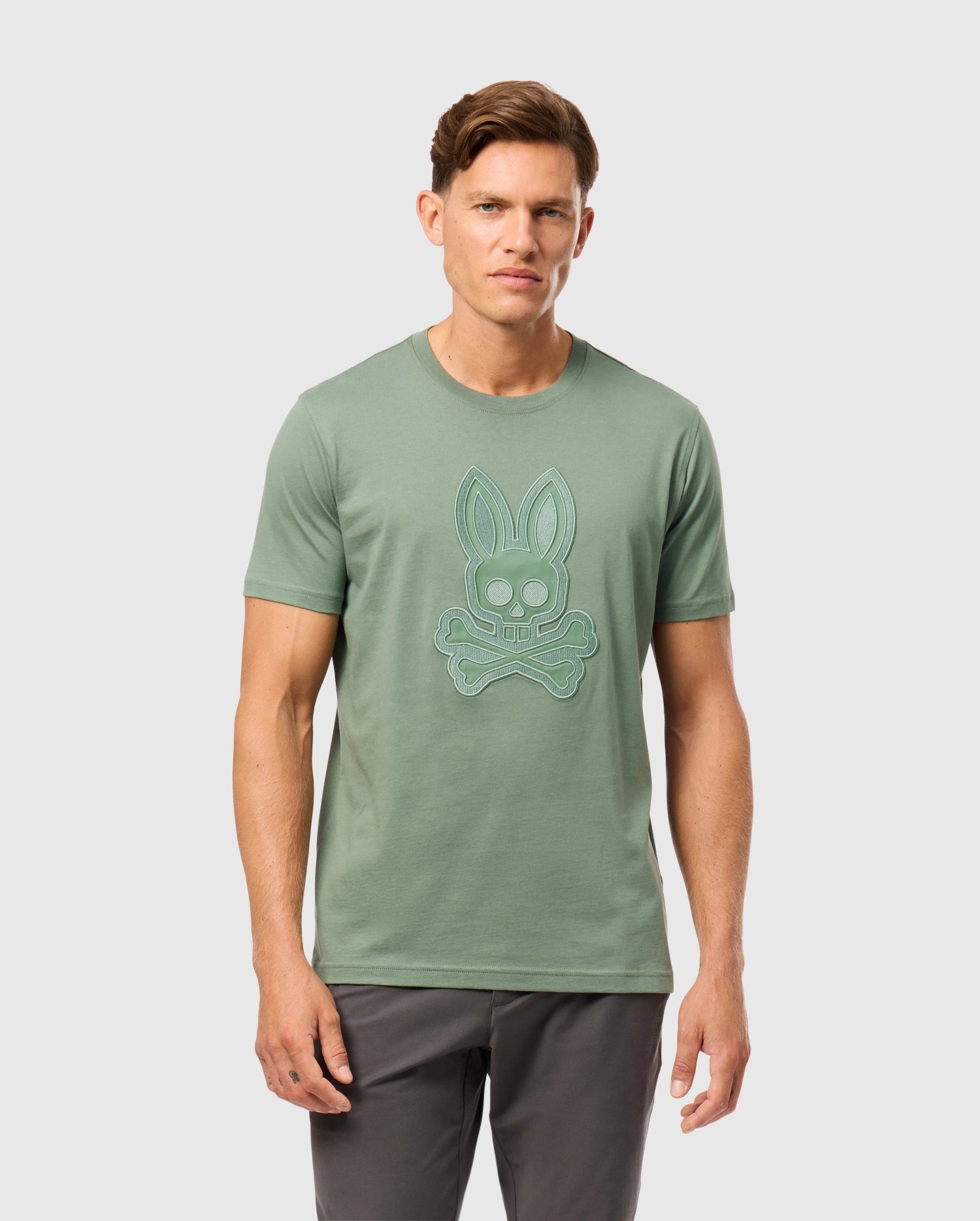 A man with brown hair is wearing a light green, Peruvian-crafted Psycho Bunny MENS MALTON GRAPHIC TEE - B6U795D200, made from Pima cotton and featuring a stylized bunny skull and crossbones design on the front. He is also wearing dark gray pants and standing against a plain white background.