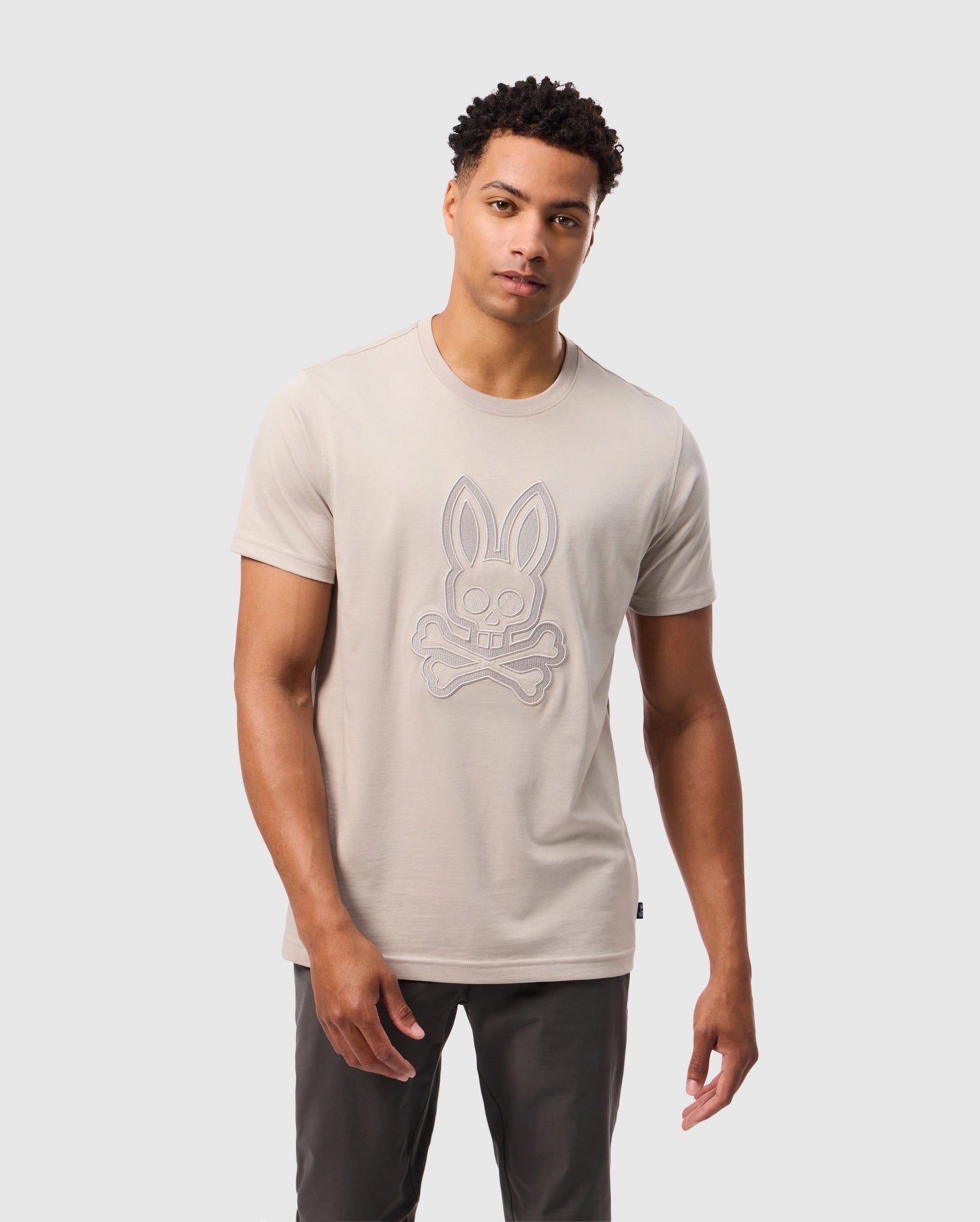 A man with short curly hair is dressed in a light gray Psycho Bunny MENS MALTON GRAPHIC TEE - B6U795D200, adorned with a bunny skull and crossbones design. Crafted from luxurious Pima cotton, he stands against a plain white background, looking at the camera with a neutral expression. He pairs it perfectly with dark trousers.