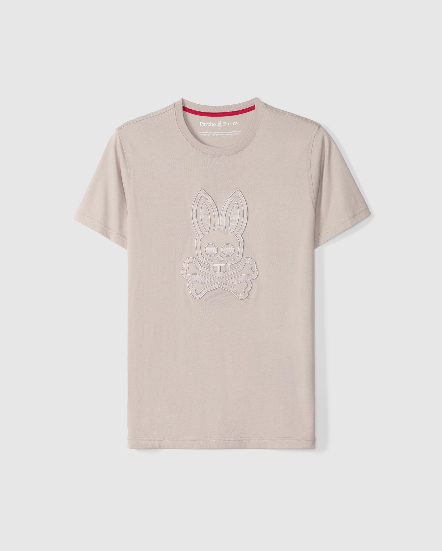 The Psycho Bunny Men's Malton Graphic Tee - B6U795D200 is a beige-colored T-shirt made from Pima cotton, featuring short sleeves and a round neck. It boasts an embossed design of a rabbit skull and crossbones. Highlighting Peruvian craftsmanship, the inner collar includes a red strip with white text. The T-shirt is showcased against a plain white background.