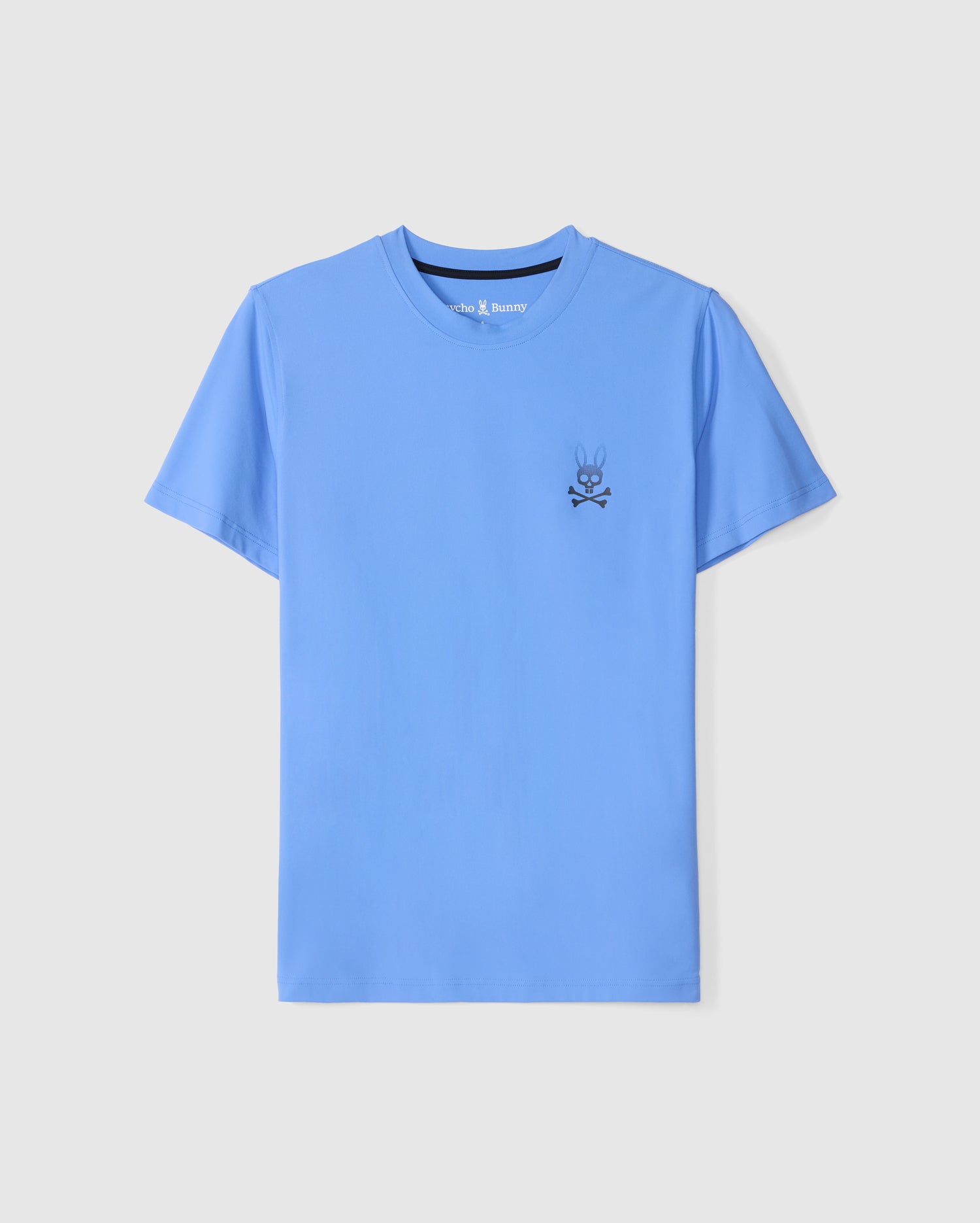 The Psycho Bunny MENS MAXWELL SPORT BACK GRAPHIC TEE - B6U799D200 is a solid light blue short-sleeve t-shirt made from breathable fabric, featuring a small black skull and crossbones graphic with bunny ears and eyes on the left chest. The shirt is displayed on a white background.