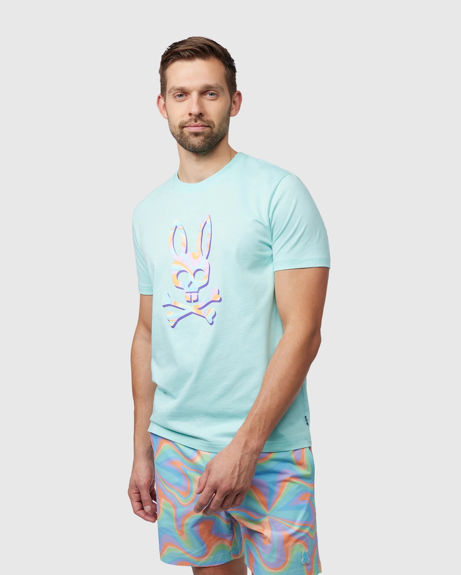 A man with short brown hair stands facing forward, wearing a light blue Psycho Bunny Pima cotton t-shirt with a bunny design and colorful space-dyed shorts. He has a subtle smile and looks directly at the camera.