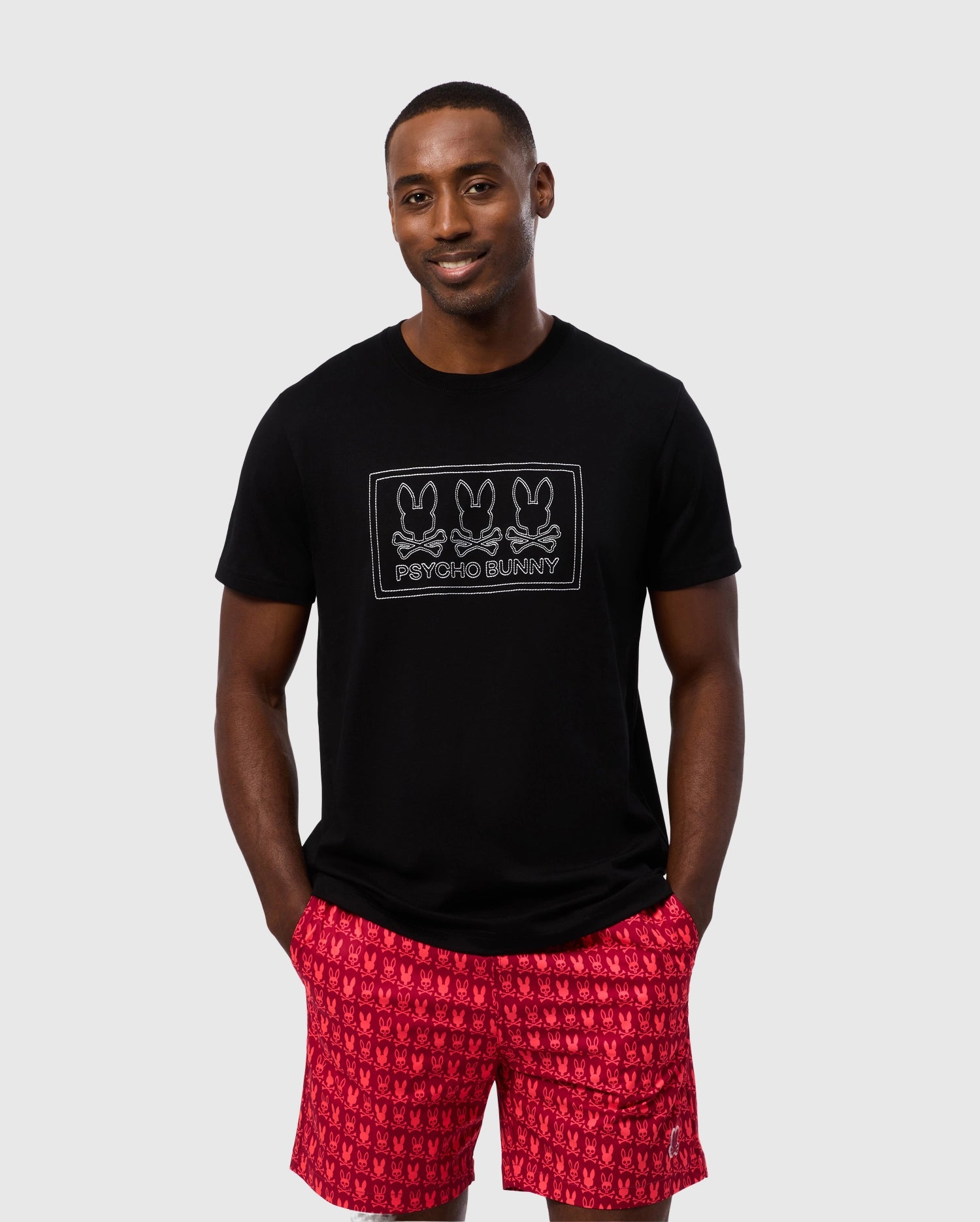 A man stands in front of a plain background wearing a black Psycho Bunny MENS TACOMA GRAPHIC TEE (B6U909D200) featuring a logo with three bunny heads and the text 