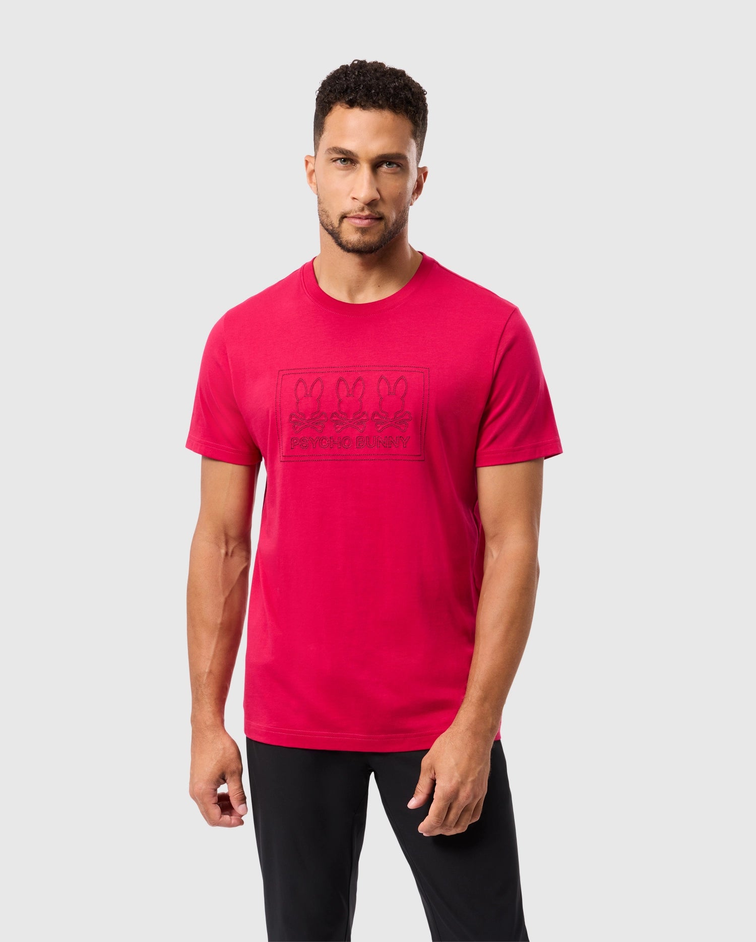 A person wearing the Psycho Bunny MENS TACOMA GRAPHIC TEE - B6U909D200 stands against a plain background. The red T-shirt, crafted from soft Pima cotton, showcases embroidered details and an embossed pattern with illustrations. The individual has short, curly hair and is looking towards the camera.