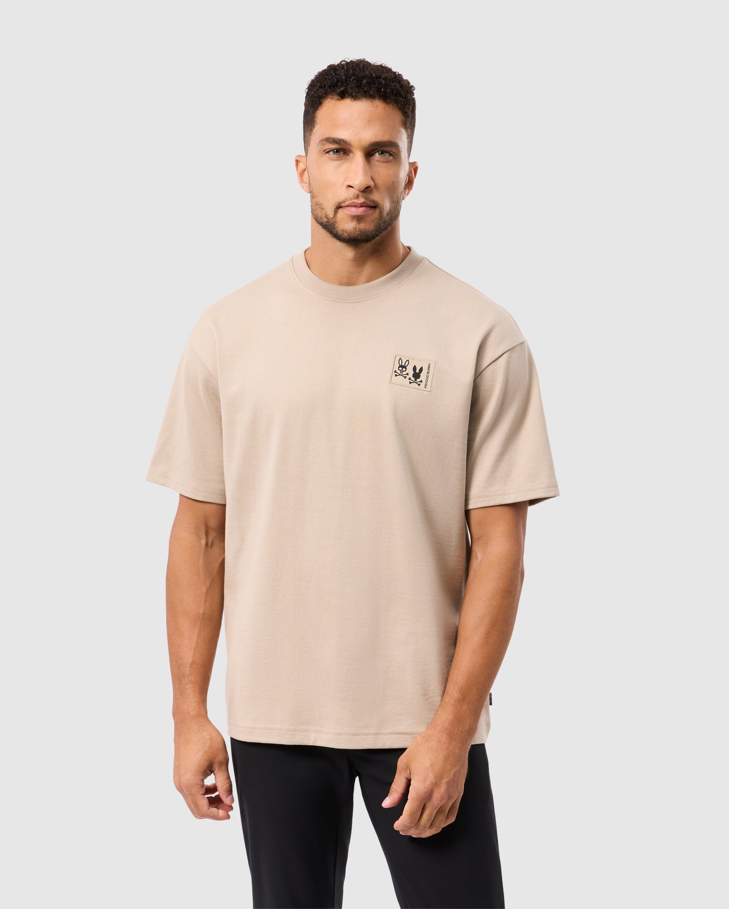 A man in a beige Psycho Bunny MENS MONROE HEAVY WEIGHT RELAXED FIT TEE (B6U910D200), made of soft Pima cotton with a chest graphic, stands against a light gray backdrop. With short dark hair, a beard, and black pants, he looks casually at the camera.