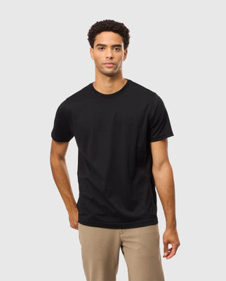 A person wearing the MENS JAMES PREMIUM CREW NECK TEE by Psycho Bunny, a regular fit black tee made of Premium Pima cotton, along with beige pants stands against a plain light gray background. They have short curly hair and a neutral expression.