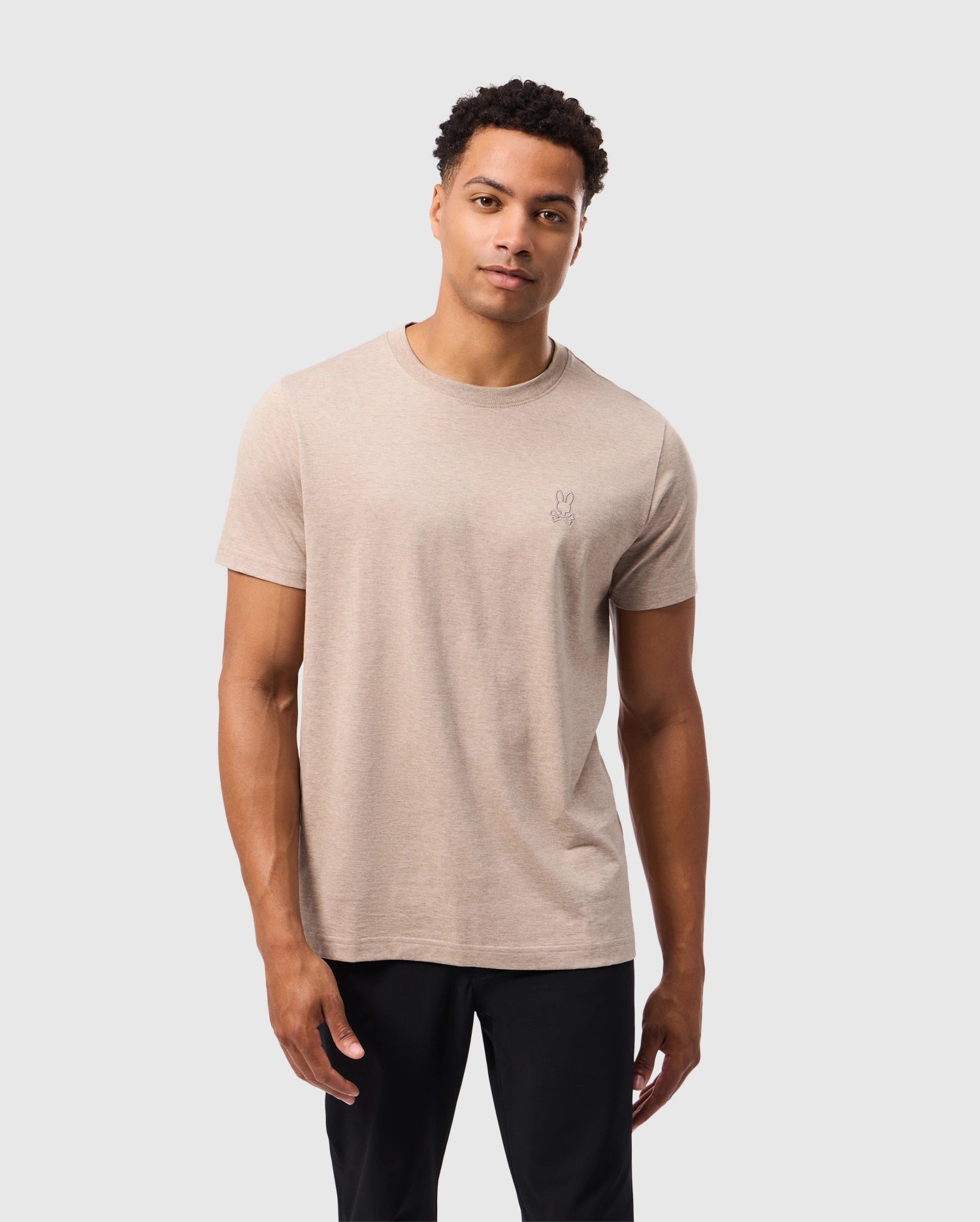 A person stands against a plain background wearing a beige Psycho Bunny MENS JAMES PREMIUM CREW NECK TEE (B6U952D200) crafted from premium Pima cotton and black pants. The t-shirt features meticulous embroidery with a small, subtle design on the chest. The individual is looking at the camera with a neutral expression, their arms relaxed by their sides.