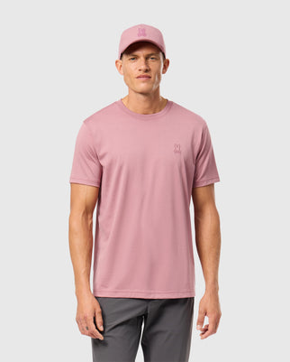 A person is wearing a pink Psycho Bunny MENS JAMES PREMIUM CREW NECK TEE - B6U952D200 and matching cap, both featuring a small embroidered rabbit logo. They are also wearing gray pants against a plain light gray background.