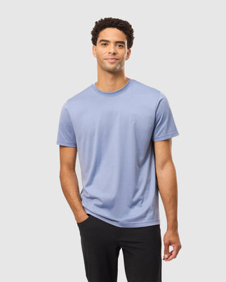 A man wearing a Psycho Bunny MENS JAMES PREMIUM CREW NECK TEE - B6U952D200, accompanied by black pants, stands against a plain white background. Crafted from premium Pima cotton, he gazes forward with a relaxed expression and one hand in his pocket.