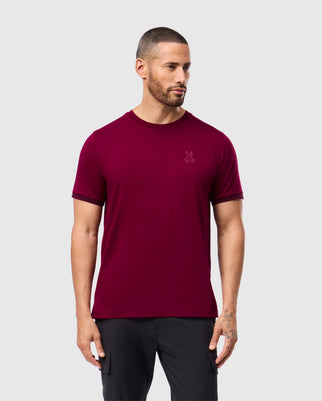 A man with short hair and a beard is wearing the stylish MENS MALTON FASHION TEE - B6U966D200 from Psycho Bunny, paired with black pants. He is standing against a plain light gray background, looking to the side with his arms by his sides, embodying modern men's fashion tee elegance.