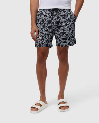 A person wearing navy blue Psycho Bunny MAYBROOK LIGHTWEIGHT SWIM TRUNKS and white slide sandals standing against a white background. Only the lower half of the body is visible.