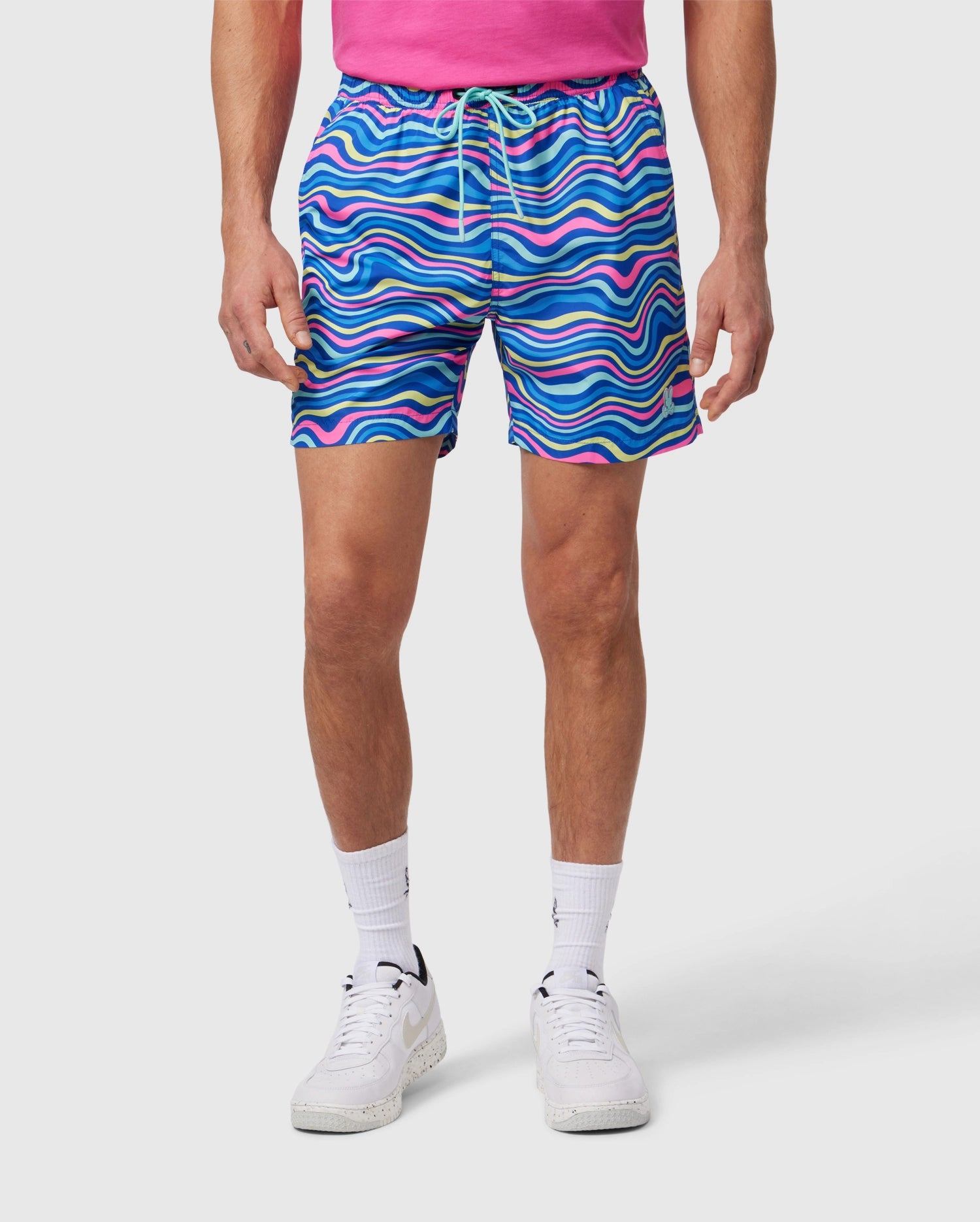 A person wearing a pink shirt, Psycho Bunny MENS CLARKSON LIGHTWEIGHT SWIM TRUNK - B6W120B2SW with a wave-like pattern in shades of blue, pink, and purple, white socks, and white sneakers stands against a plain white background. The photo is cropped to show from the neck down to the feet.