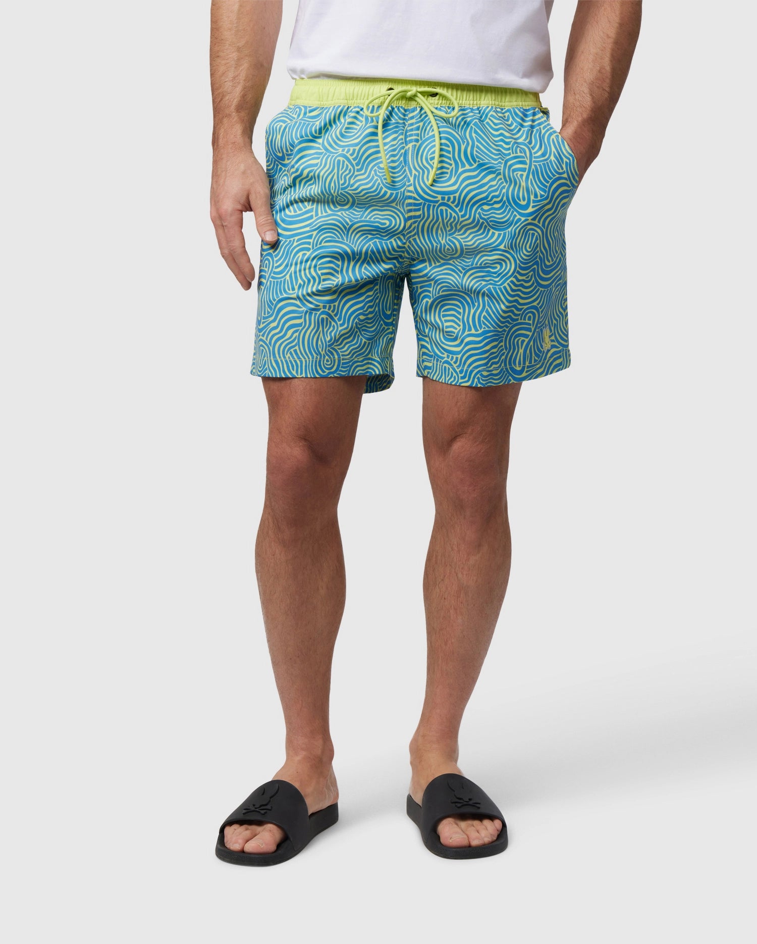 Man wearing a white t-shirt, VERONA PRINT SWIM TRUNK - B6W172B2SW from Psycho Bunny with a drawstring, and black slide sandals. The shorts, with an abstract wavy design in blue and yellow, are made from quick-dry fabric. He stands against a plain white background with his hands in his pockets.