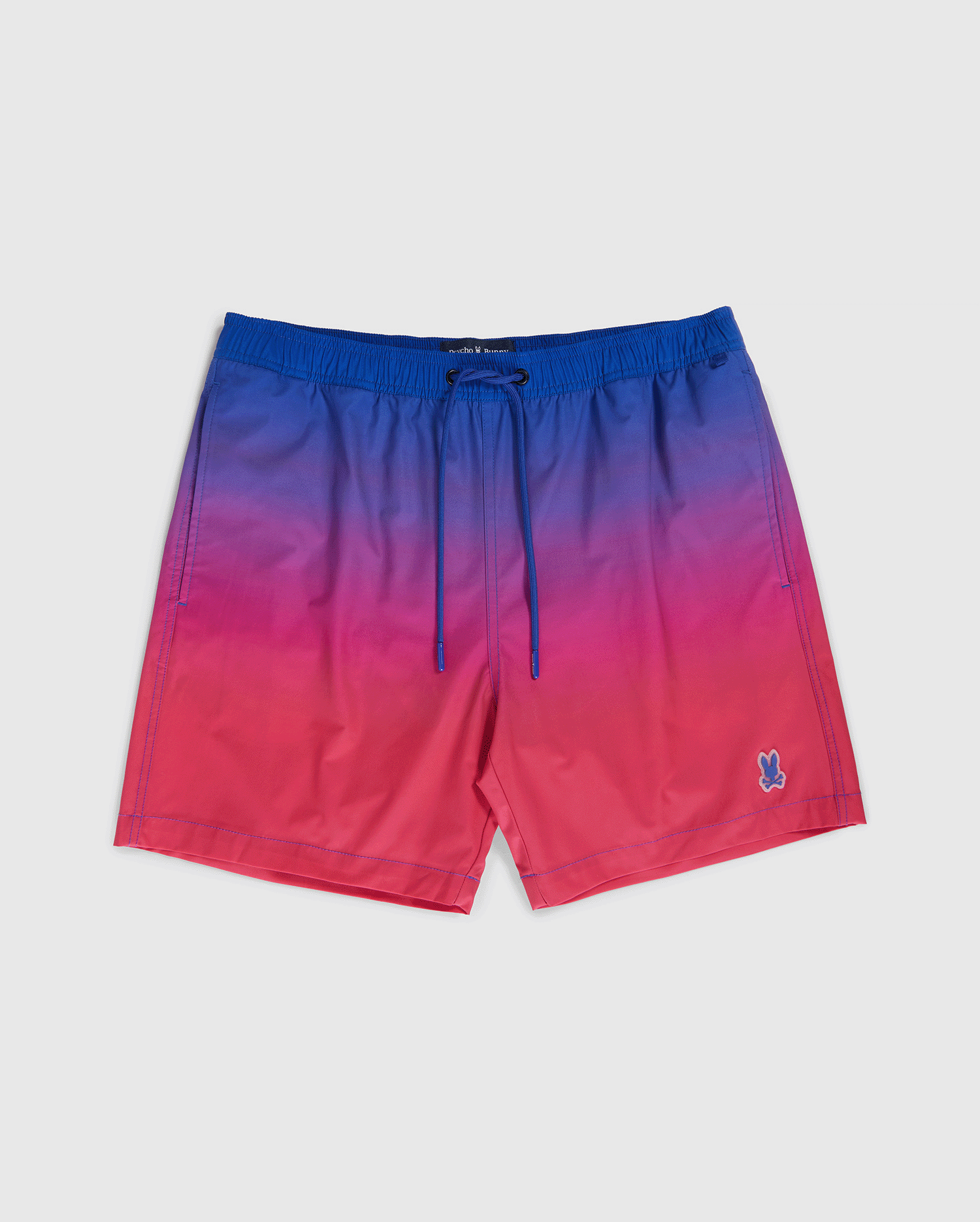 A pair of Psycho Bunny MENS MALTA HYDROCHROMIC SWIM TRUNK - B6W173B2SW with an ombré design that transitions from blue at the top to pink at the bottom. They feature an elastic waistband with a drawstring and a small embroidered Bunny pattern on the left leg.