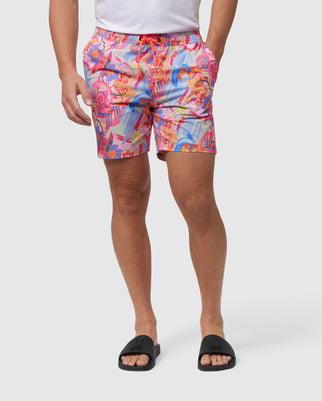 A person is wearing Psycho Bunny MENS BENTON SWIM TRUNK - B6W319B2SW featuring a colorful, graphic print with a mix of pink, blue, and orange abstract designs. The individual is also wearing black slide sandals. The background is plain and white, focusing on the lower half of the body.