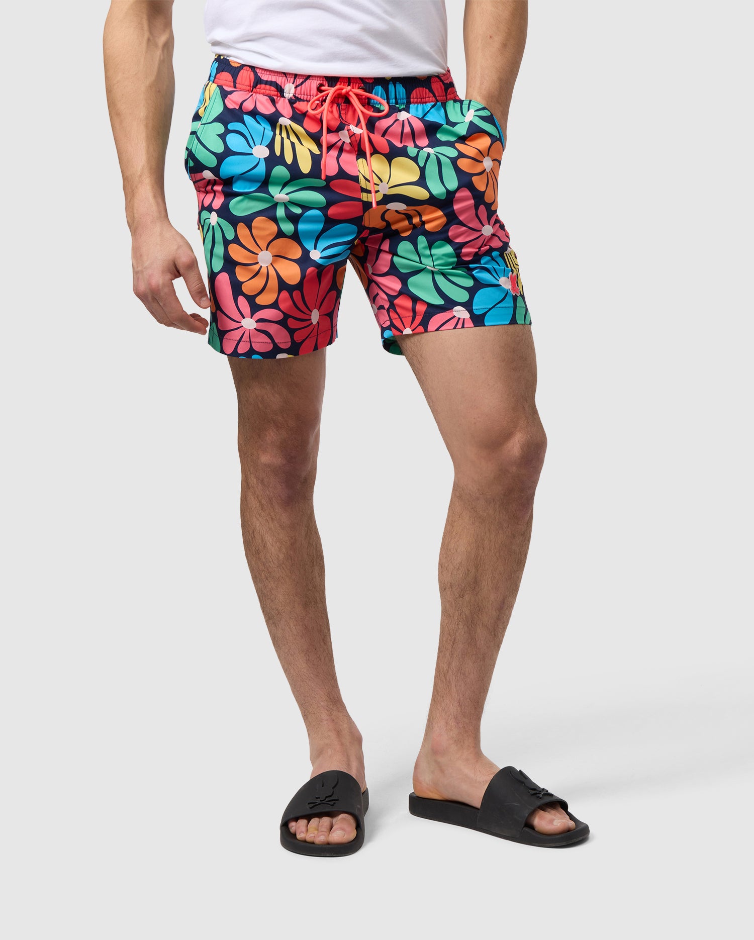 A person is standing, wearing Psycho Bunny's MENS MENTZ SWIM TRUNK - B6W320B2SW with a vibrant floral pattern and black slides. The swim trunks have a bold red drawstring, and the individual's hands are hanging relaxed at their sides. A waterproof bag rests nearby against the plain, light-colored background.