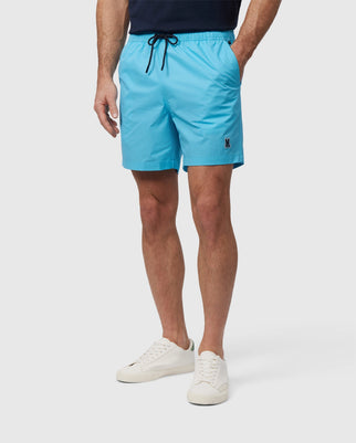 A man is shown from the waist down, wearing light blue Psycho Bunny MENS MALTA HYDROCHROMIC SWIM TRUNK and white sneakers, posing against a plain background. The shorts have an embroidered logo on the left thigh.