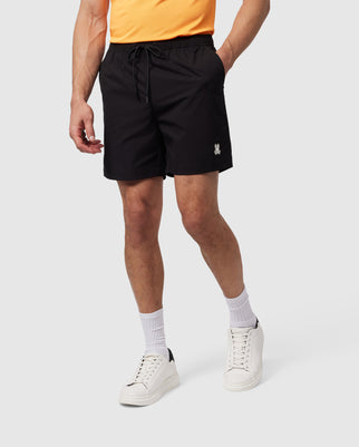 A man wearing a bright orange shirt, black mechanical stretch shorts, white socks, and white sneakers stands with his hand resting on his thigh, against a neutral background in Psycho Bunny's MENS MALTA HYDROCHROMIC SWIM TRUNK - B6W321B2SW.