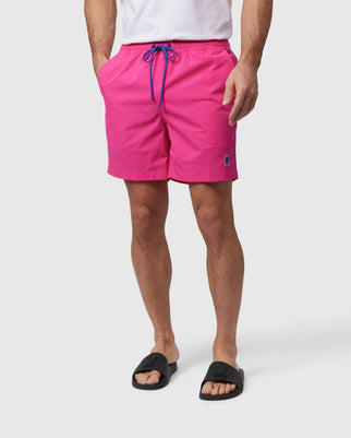 A person is wearing a white t-shirt paired with bright pink Psycho Bunny MENS MALTA HYDROCHROMIC SWIM TRUNK - B6W321B2SW that have a blue drawstring and a small embroidered Bunny pattern. The person is also wearing black slide sandals with a minimalist design. The background is plain and white.