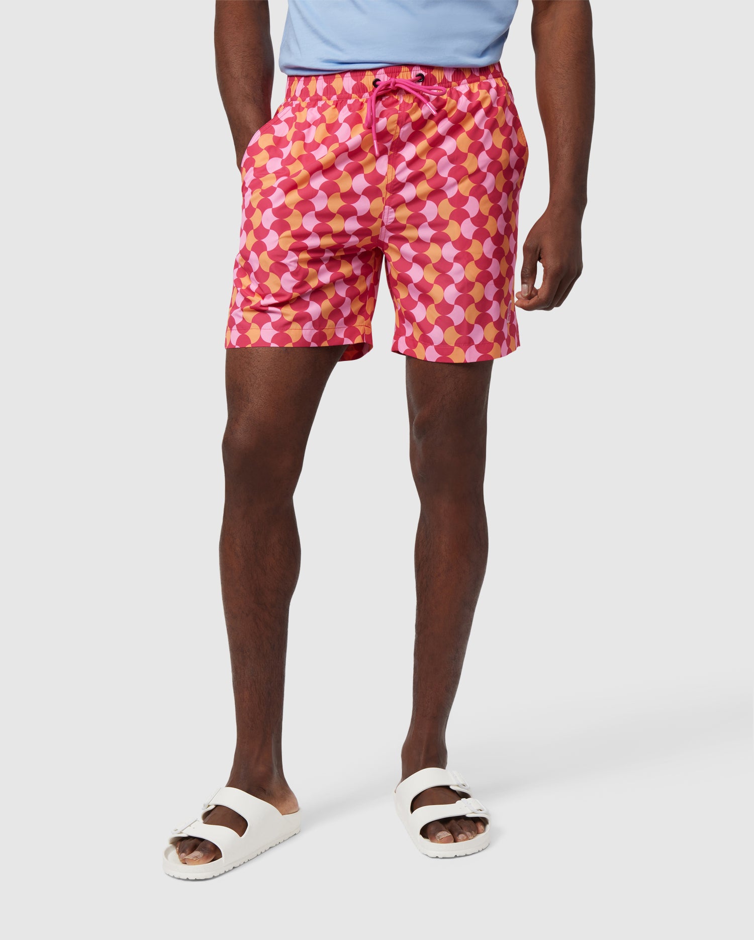 A person is wearing a light blue shirt tucked into Psycho Bunny's MENS COLCHESTER LIGHTWEIGHT SWIM TRUNK - B6W348B2SW, which features a vibrant graphic pattern and is made from recycled polyester, with hands in the pockets. They are also wearing white slide sandals. The background is plain and white.