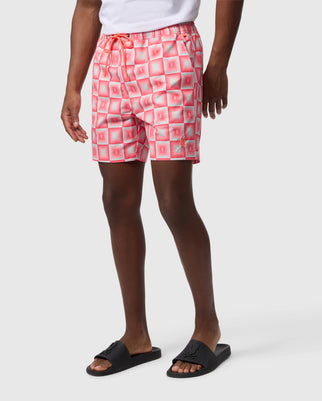 A person wears patterned pink and white Psycho Bunny AOP swim trunks and black slide sandals, shown from waist to calves with a plain white background.