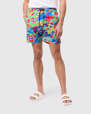 A person is standing with hands in the pockets of their brightly colored, checkered Psycho Bunny MENS DENNIS ALL OVER PRINT SWIM TRUNK - B6W530C200 shorts. The swim trunks feature a vibrant blend of red, blue, green, and yellow quick-dry fabric. They are also wearing white slide sandals. The background is plain light gray.